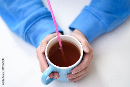 Kubek z gorącą herbatą trzymany w dłoniach, różowa plastikowa słomka jednorazowego użytku w środku  photo