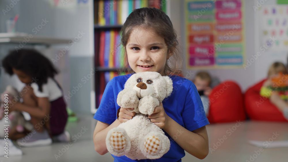 Close up of little girl with teddy bear smiling at camera sitting on floor in kindergarten playroom Close up of little girl with teddy bear smiling at camera sitting on floor in kindergarten playroom