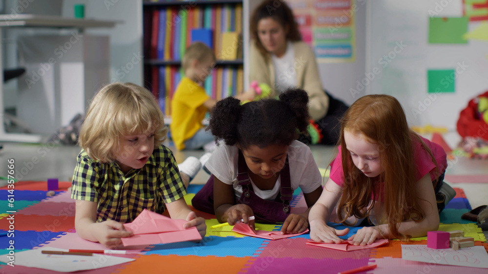 Fototapeta premium Little kids learn to folding paper origami lying on floor in kindergarten