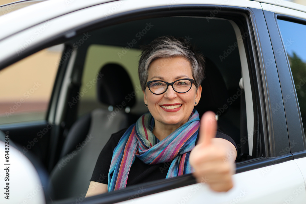 Cheerful senior woman in glasses sits in the driver's seat and shows thumbs up.