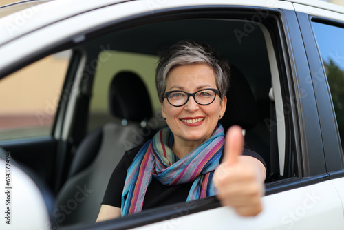 Cheerful senior woman in glasses sits in the driver\'s seat and shows thumbs up.