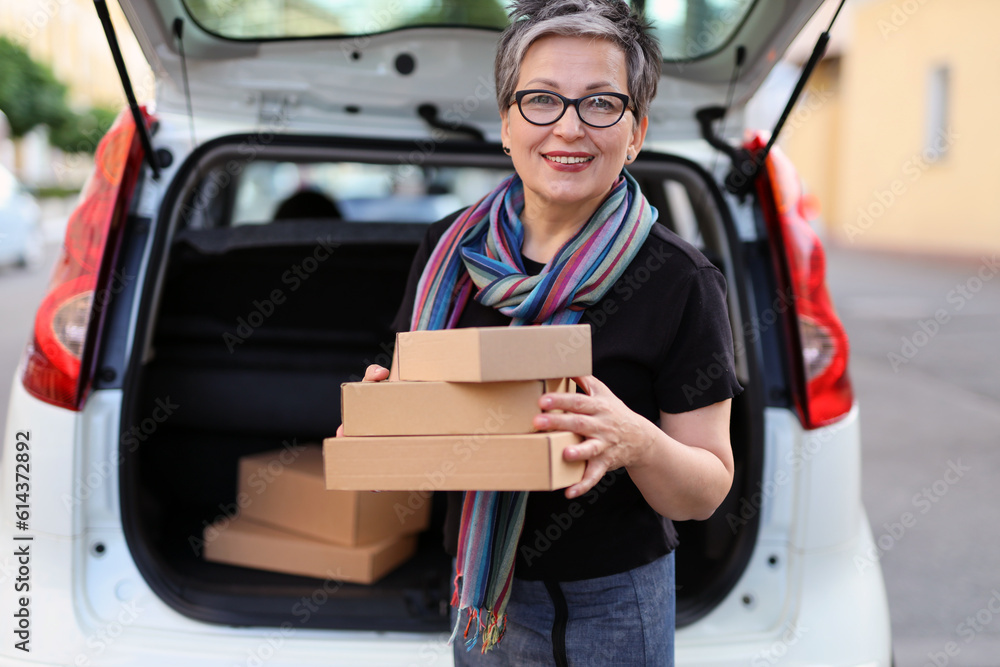 Smiling mature woman in glasses with boxes in her hands is loading parcels into the trunk of a car. Smiling mature woman in glasses with boxes in her hands is loading parcels into the trunk of a car.