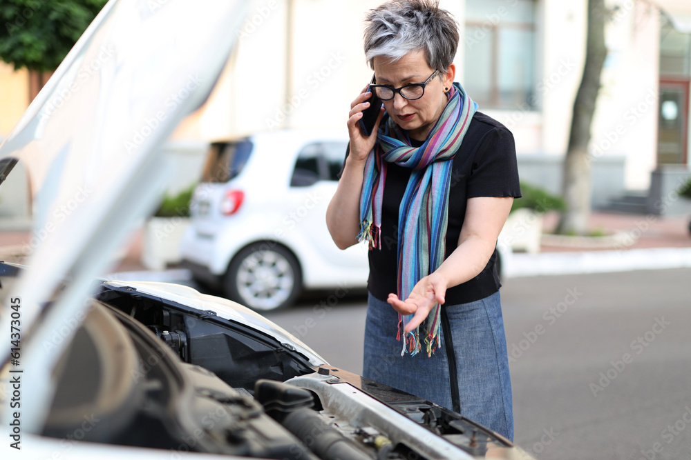 Perplexed mature woman talking on a smartphone against the background of an open car hood.