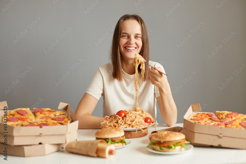 Attractive smiling woman with brown hair wearing white T-shirt sitting at table among unhealthy food isolated over gray background enjoying pasta at home eating junk dish.