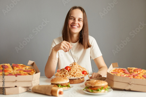 Extremely happy smiling beautiful woman with brown hair wearing white T-shirt sitting at table among fast food isolated over gray background enjoying unhealthy nutrition.