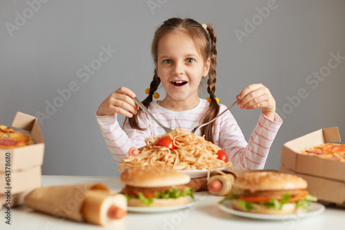 Amazed excited little girl with braids sitting at table with junk food isolated over gray background holding forks eating pasta looking at camera with big eyes.