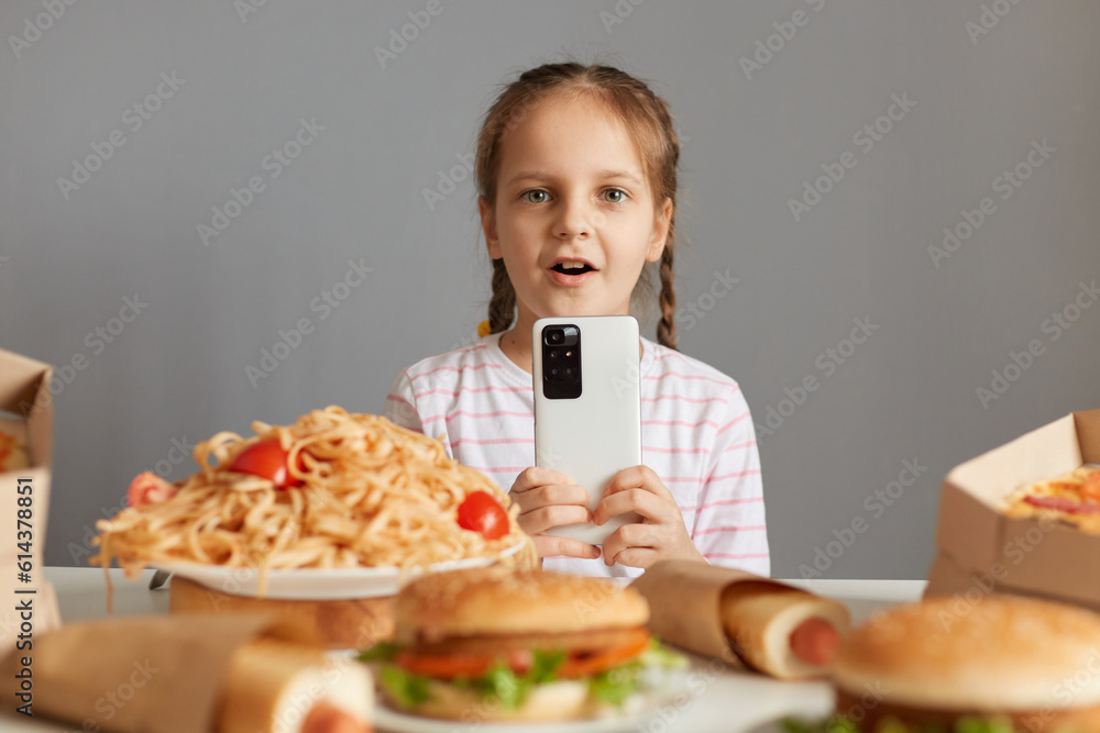 Shocked astonished little girl with braids sitting at table with junk food isolated over gray background looking at camera with big eyes uaing smartp phone online food delivery. Shocked astonished little girl with braids sitting at table with junk food isolated over gray background looking at camera with big eyes uaing smartp phone online food delivery.