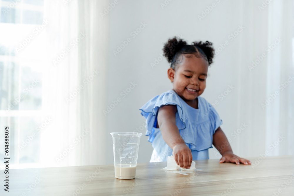 A cute little girl enjoys cleaning spilled milk on the table. Focus on a glass of milk. A cute little girl enjoys cleaning spilled milk on the table. Focus on a glass of milk.
