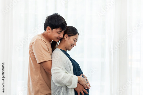 Close up - Smiling Asian man wearing a brown shirt hugs a pregnant woman in blue. White curtain background.