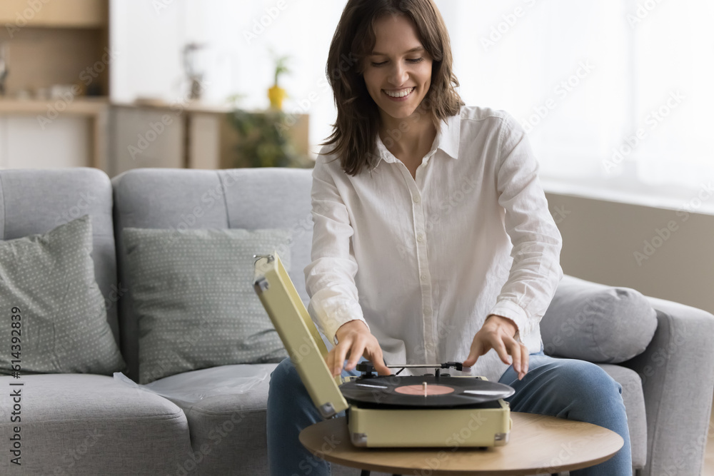 Happy woman placing vinyl record on turntable seated on couch, spend pleasant leisure resting at home, play nostalgic music, enjoy favourite melody, tune from the past. Retro lover pastime and hobby Happy woman placing vinyl record on turntable seated on couch, spend pleasant leisure resting at home, play nostalgic music, enjoy favourite melody, tune from the past. Retro lover pastime and hobby