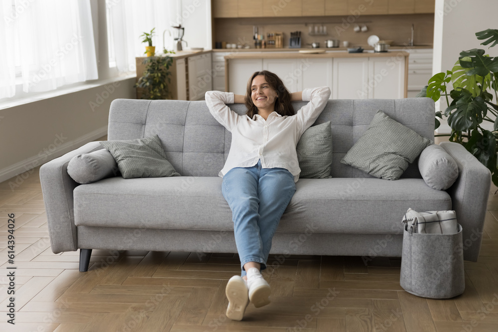 Millennial woman in casual clothes lying down on sofa with hands behind head smile feel happy on weekend at smart home with climate control enjoy cool air in summer day in studio apartment. Relaxation - obrazy, fototapety, plakaty 