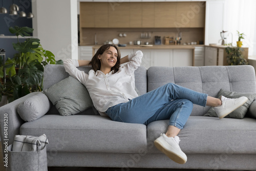 Fototapeta Cheerful millennial woman relax on sofa in studio apartment, leaned on soft comf