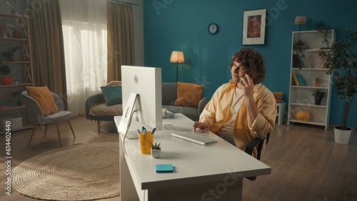 A young curly guy is sitting at a computer with a smartphone to his ear. A man is talking on the phone while working on the computer. The concept of technology, communication, connection.
