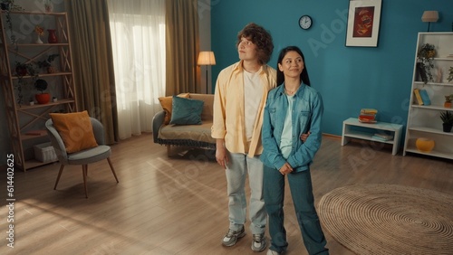 A young couple is standing in a cozy living room and examining it. The curlyhaired young man put his arm around the girl's waist. Young people admire their family nest.