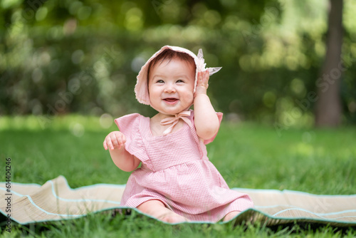 adorable and happy baby girl outdoors in the park - portrait of 7 or 8 months old beautiful little child smiling cheerful sitting on mat on grass at city park wearing cozy hat and dress