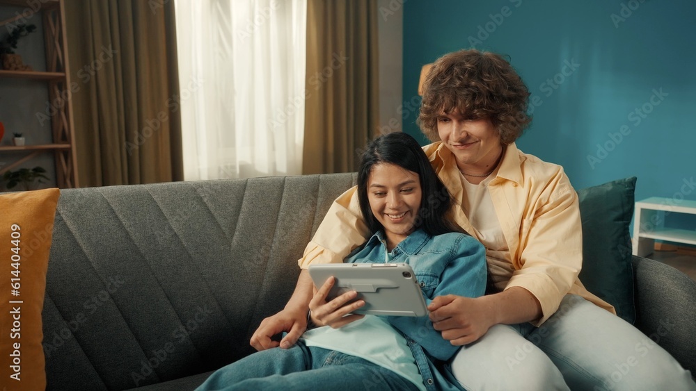 A couple spends time together on a day off. a Woman and a man watch a movie, photo, video on a tablet. A Asian woman lies on a sofa with a tablet in her hands, leaning her back on a man. Close up. A couple spends time together on a day off. a Woman and a man watch a movie, photo, video on a tablet. A Asian woman lies on a sofa with a tablet in her hands, leaning her back on a man. Close up.