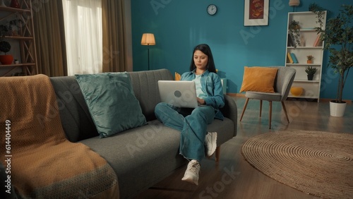Attractive Asian woman is sitting on the sofa in the living room with a laptop on her lap. A woman works, browses the Internet, looks at photos, videos. Remote work, freelancer.