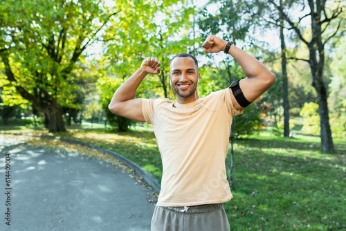 Portrait successful sportsman man satisfied with training results and achievements  looking at camera and holding hands up in triumph gesture  hispanic man running in park with headphones and phone.