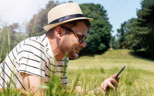 Side view man wearing straw hat and sunglasses lying on front on green grass of lawn and using his smart phone and smiling photo