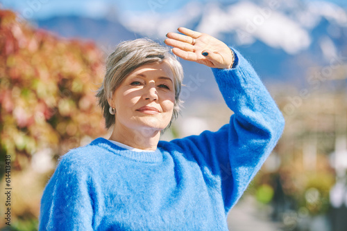 Outdoor portrait of beautiful mature 55 - 60 year old woman, wearing blue pullover, hiding face from the sun with hand