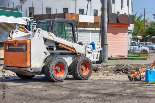 A highly manoeuvrable construction vehicle with a jackhammer attached is parked at a work site with a city street being repaired in the background. photo