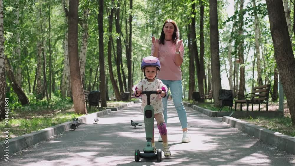 Mom teaches daughter to ride a scooter in the park in summer. Mom teaches daughter to ride a scooter in the park in summer.