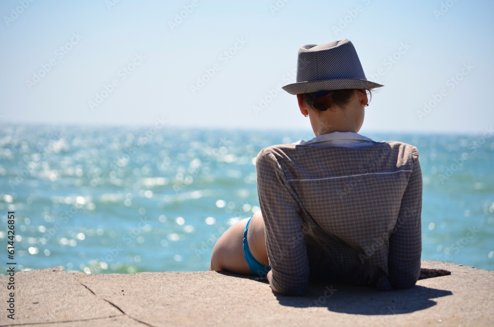 A young girl in a hat is sunbathing on the pier and looking at the sea. View from the back.