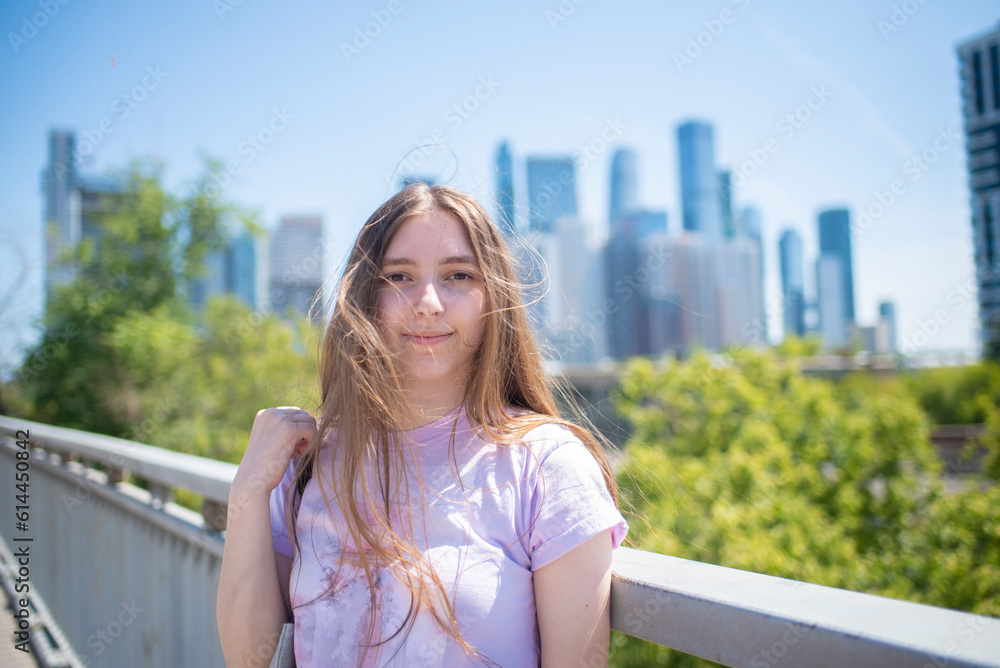 portrait of a smiling teenager girl, a student with very long hair, stands on a bridge in front of skyscrapers