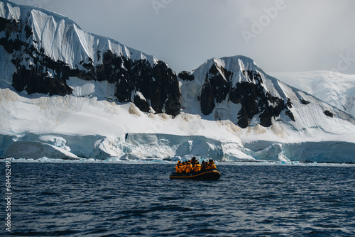 Antarctic expedition, cruise passengers in yellow parkas ride in a Zodiac inflatable boat, very close to a huge white iceberg. Climate change and global warming.