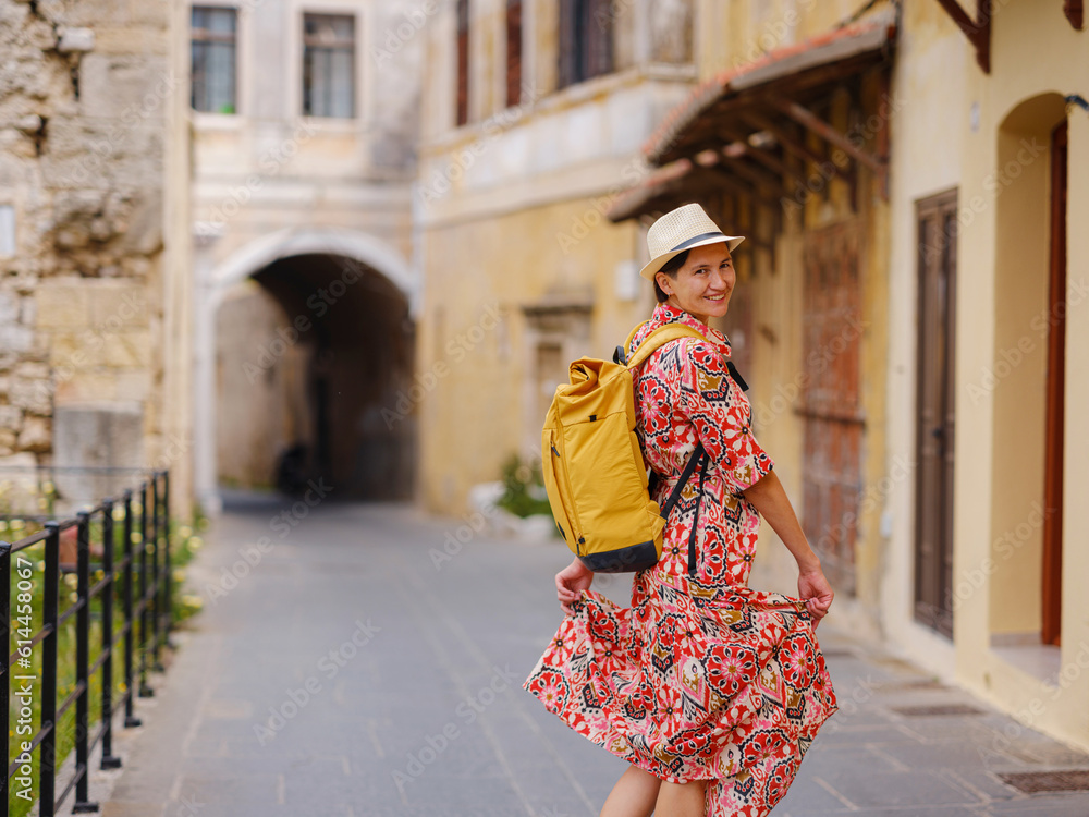 Young Asian woman in red dress and backpack walks and looks at cozy narrow streets of old city. Tourism, vacation, and discovery concept, female traveler visiting southern Europe, Rhodes island Greece