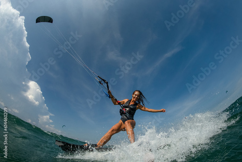 Kitesurfing girl in black sexy swimsuit with kite in sky on board in blue sea riding waves with water splash. Recreational activity, water sports, action, hobby and fun in summer time. 