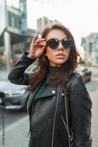 Beautiful fashion young stylish woman model in trendy clothes with a green dress and a fashionable leather black jacket walks in the city and puts on sunglasses