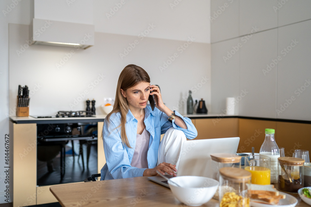 Young woman housewife sitting at kitchen table with breakfast speaking on mobile phone while paying household bills online. Female freelancer making business call while working remotely from home