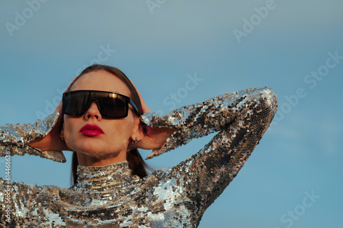 Fashionable confident woman wearing trendy black sunglasses, sequin  turtleneck top, posing outdoor, against blue sky. Close up fashion portrait. Copy, empty space for text