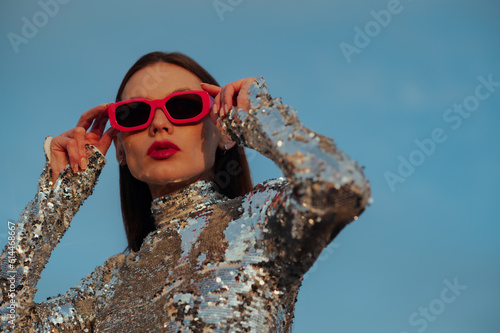 Fotografering Fashionable confident woman wearing trendy fuchsia color rectangular sunglasses, sequin  turtleneck top, posing outdoor, against blue sky