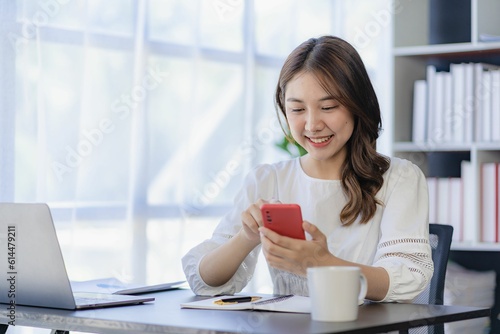 Smiling Asian woman talking on the phone with a customer Young positive female accountant using smartphone talking to team at her desk relaxing with mobile application