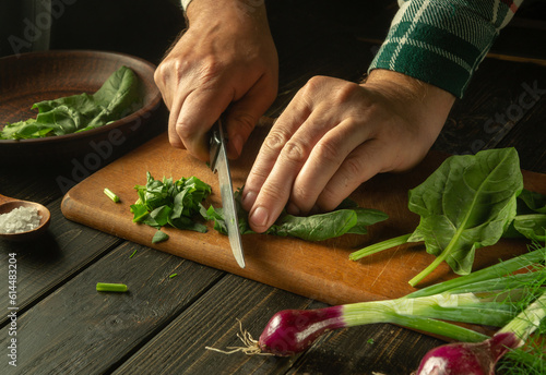 Slicing spinach leaves for a diet salad for lunch. The hands of the cook are preparing a salad of spinach and fresh vegetables on a kitchen board