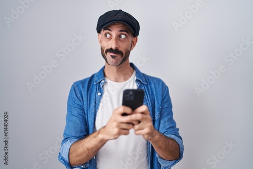 Hispanic man with beard using smartphone typing message smiling looking to the side and staring away thinking. © Krakenimages.com