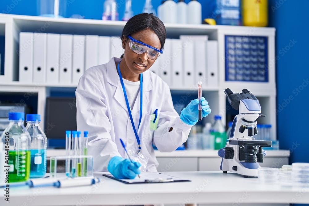 Young african american woman scientist writing on document holding blood test tube at laboratory