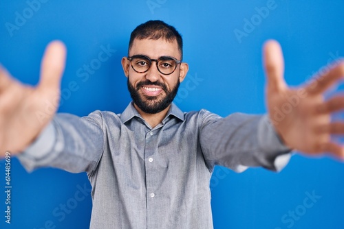 Middle east man with beard standing over blue background looking at the camera smiling with open arms for hug. cheerful expression embracing happiness.