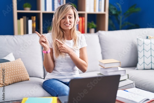 Young blonde woman studying using computer laptop at home pointing aside worried and nervous with both hands, concerned and surprised expression