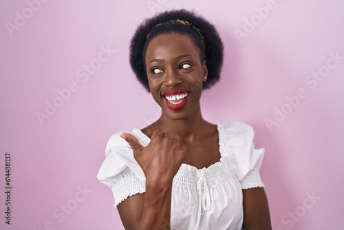 African woman with curly hair standing over pink background smiling with happy face looking and pointing to the side with thumb up.