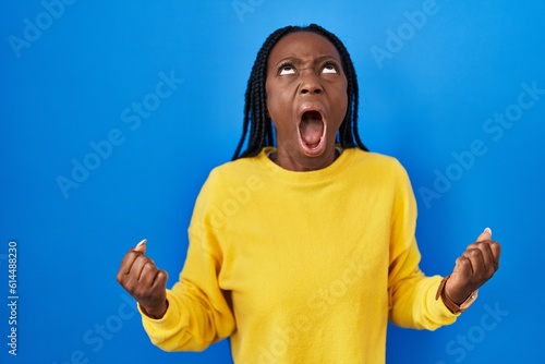 Beautiful black woman standing over blue background angry and mad screaming frustrated and furious, shouting with anger. rage and aggressive concept.