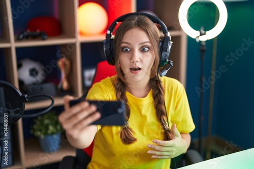 Young caucasian woman playing video games with smartphone scared and amazed with open mouth for surprise, disbelief face