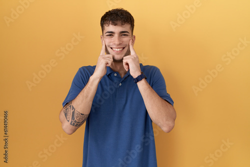 Young hispanic man standing over yellow background smiling with open mouth, fingers pointing and forcing cheerful smile