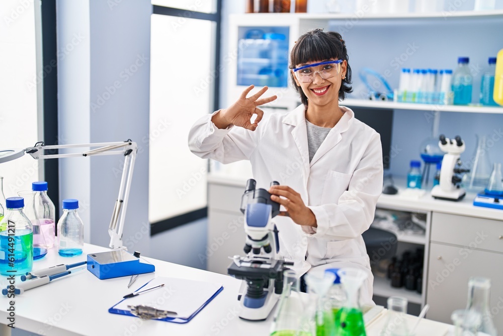 Young hispanic woman working at scientist laboratory doing ok sign with fingers, smiling friendly gesturing excellent symbol Young hispanic woman working at scientist laboratory doing ok sign with fingers, smiling friendly gesturing excellent symbol
