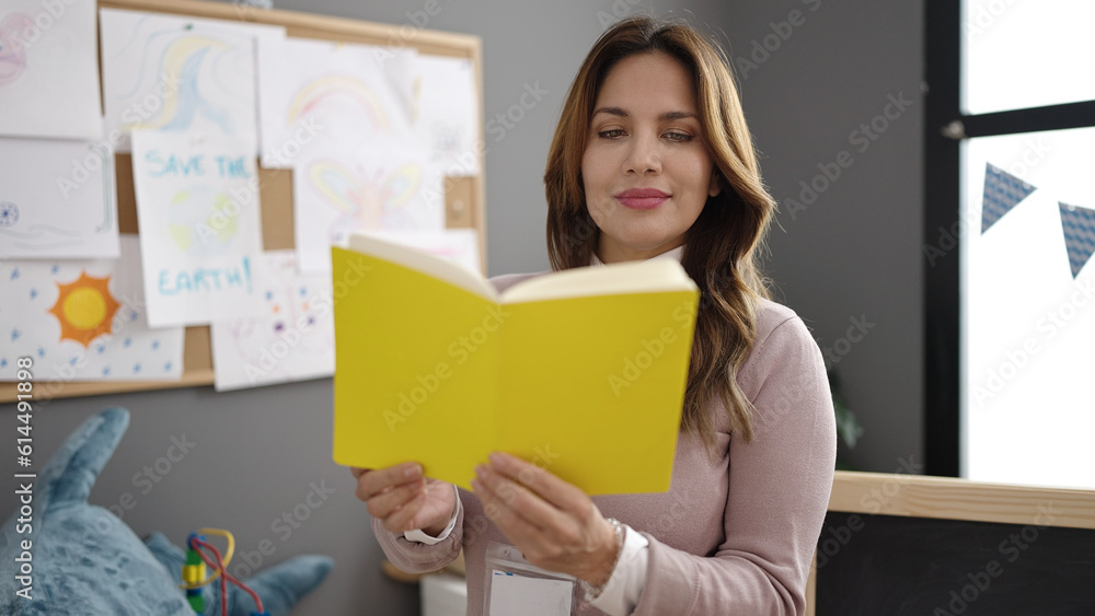 Young beautiful hispanic woman preschool teacher reading book standing at kindergarten Young beautiful hispanic woman preschool teacher reading book standing at kindergarten