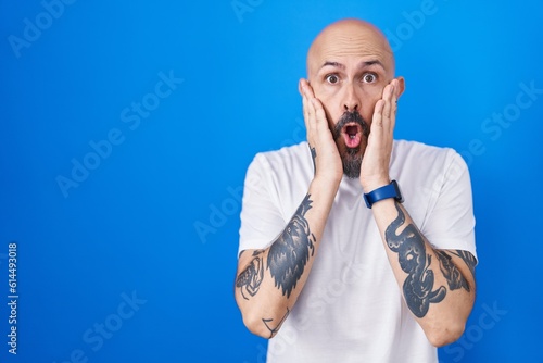 Hispanic man with tattoos standing over blue background afraid and shocked, surprise and amazed expression with hands on face