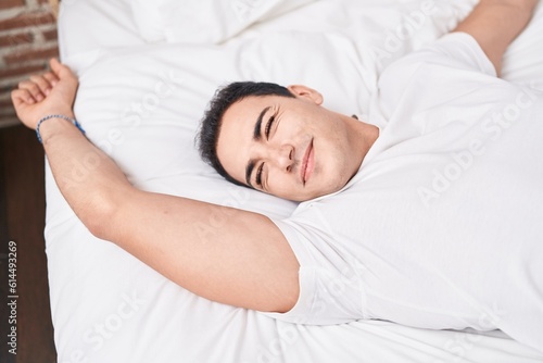 Young hispanic man waking up stretching arms at bedroom