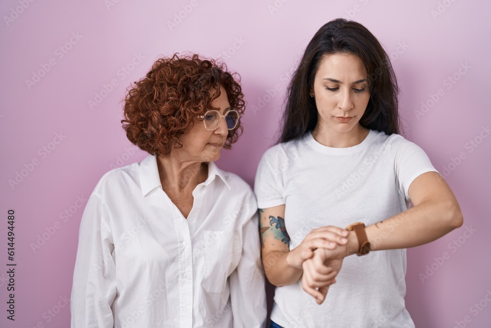 Hispanic mother and daughter wearing casual white t shirt over pink background checking the time on wrist watch, relaxed and confident